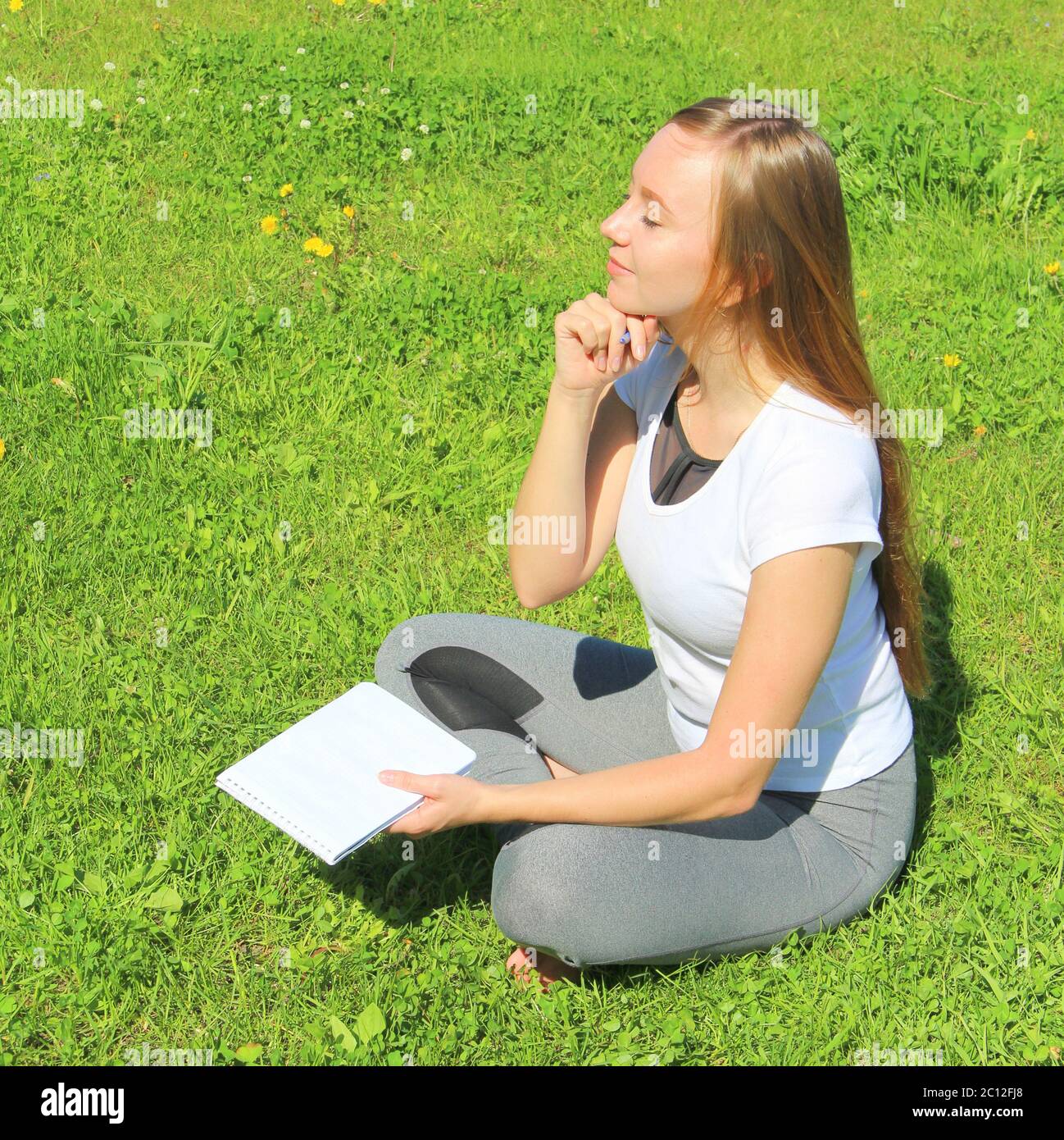 Ein junges, schönes Mädchen im weißen T-Shirt mit geschlossenen Augen sitzt auf dem grünen Gras auf dem Rasen mit einem Notizbuch und einem Stift, mit dem Handkinn gestützt, denkt und träumt, komponiert Text und Gedichte. Stockfoto
