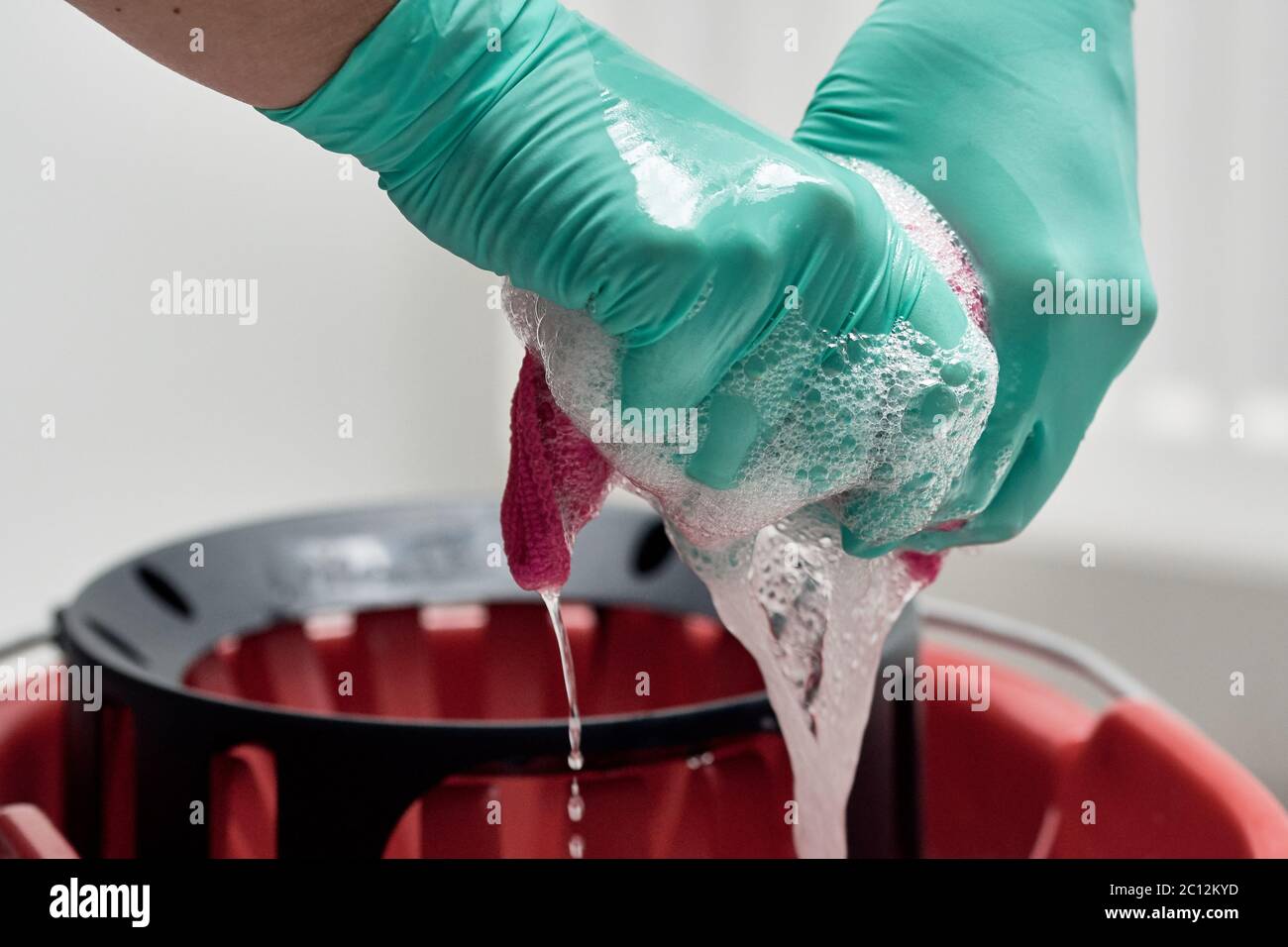 Nahaufnahme der Hände mit Handschuhen, die ein Reinigungstuch über den mit Wasser und Seife gefüllten Eimer auswringen Stockfoto