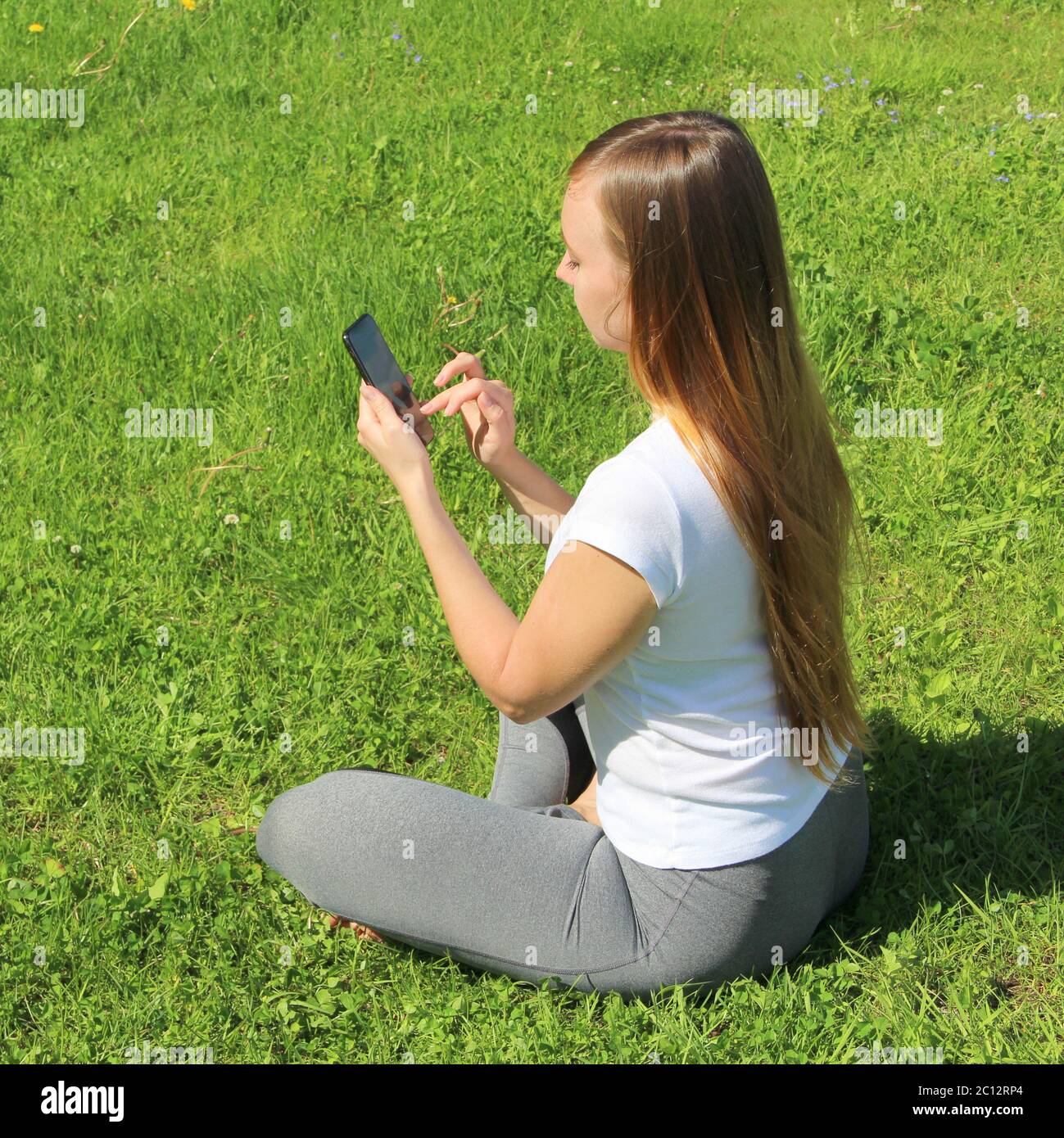 Ein junges schönes Mädchen von europäischem Aussehen in einem weißen T-Shirt mit langen blonden Haaren sitzt auf grünem Gras, auf dem Rasen mit einem Handy in den Händen, kippt durch soziale Netzwerke, arbeitet im Telefon. Stockfoto