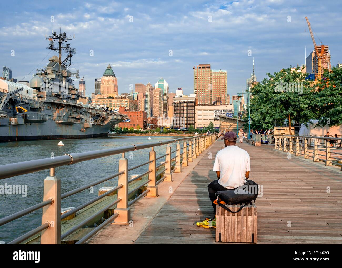 Ein nicht identifizierter Mann sitzt am Abend auf einer Bank am Pier 84 Dock. Die Skyline von Midtown Manhattan und die USS Intrepidare im Hintergrund. Stockfoto