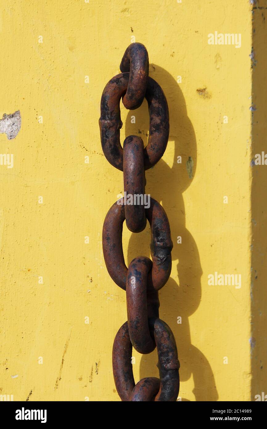 Rostige Metallkette hängt auf gelben Betonsockel und wirft einen Schatten. Stockfoto