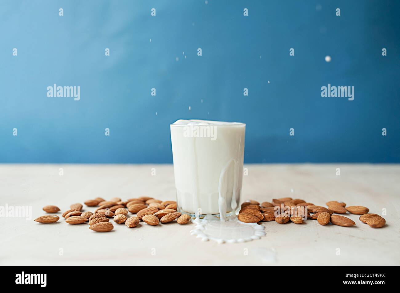 Spritzer und Tropfen aus einem klaren Becher voller Milch mit schön angereisteten Mandeln. Mandelmilch auf blauem Hintergrund Stockfoto