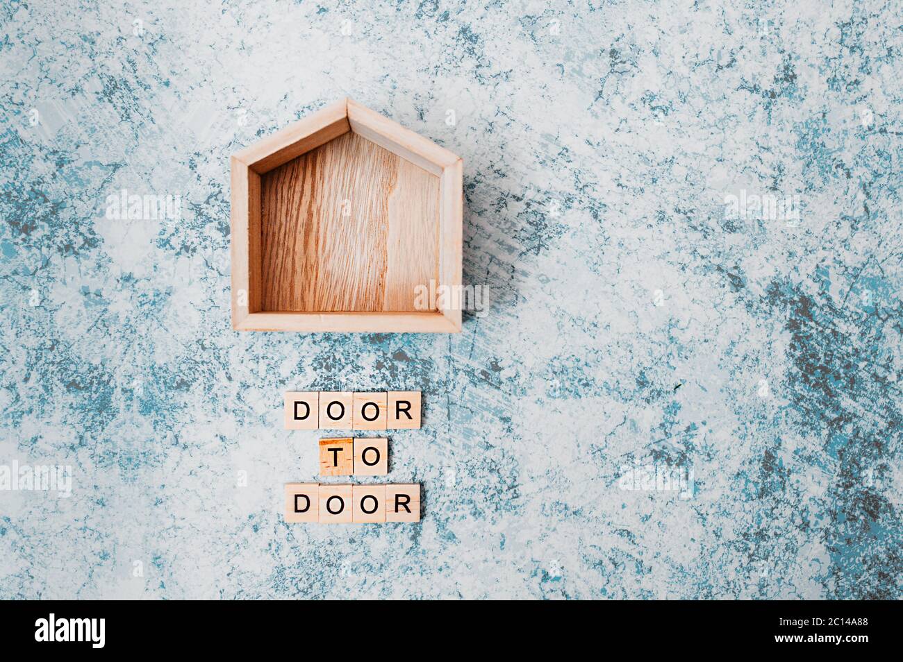 Holzdekor kleines Haus mit der Aufschrift Tür zu Tür aus hölzernen Buchstaben auf grau-blauem Zementhintergrund. Kontaktlose Lieferung und soziale distanci Stockfoto