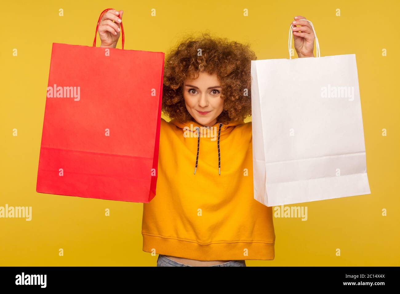 Portrait von glücklichen Shopper, lockig-haired Hipster Mädchen in Hoodie Anhebung Einkaufstaschen mit leerem Bereich, kopieren Platz auf Verpackungen für Werbetext. ind Stockfoto