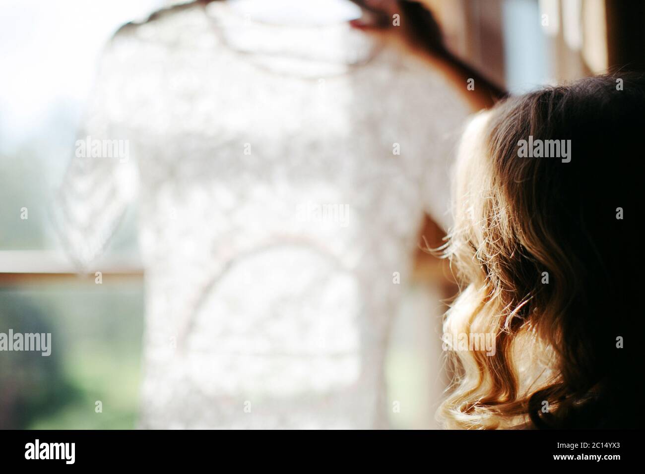 Zugeschnittene Aufnahme der blonden Frau mit lockigen Haaren halten und Blick auf schöne weiße Hochzeitskleid auf einem Kleiderbügel, in der Nähe des Fensters in ihrem Haus, b Stockfoto