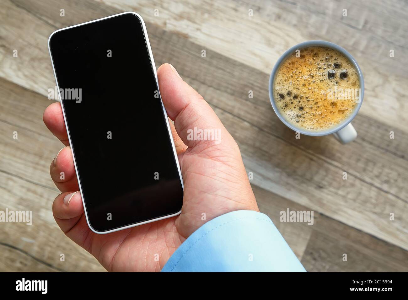Smartphone in der Hand bei der Arbeit mit einem leeren Bereich, um ein benutzerdefiniertes Bild einzufügen. Tasse Tee und Holzboden im Hintergrund Stockfoto
