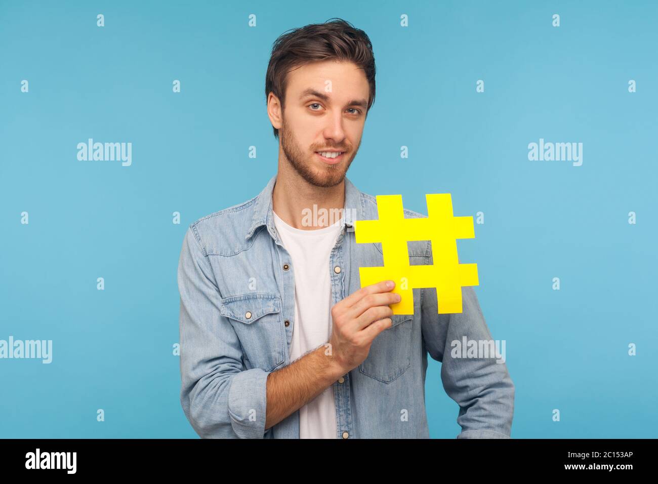 Portrait von fröhlichen Mann in Denim-Shirt mit gelben Hashtag-Symbol, isoliert auf blauem Hintergrund mit Kopierraum für getaggte Nachricht, Popula Stockfoto