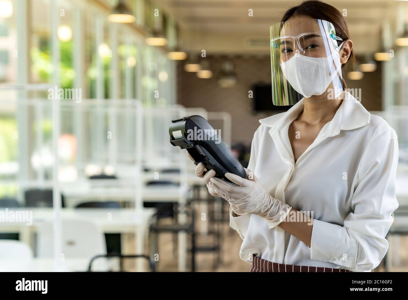Portrait Attraktive asiatische Kellnerin tragen Gesichtsmaske und Gesichtsschutz halten Kreditkartenleser für kontaktlose Zahlung mit Restaurant innen Hintergrund. N Stockfoto