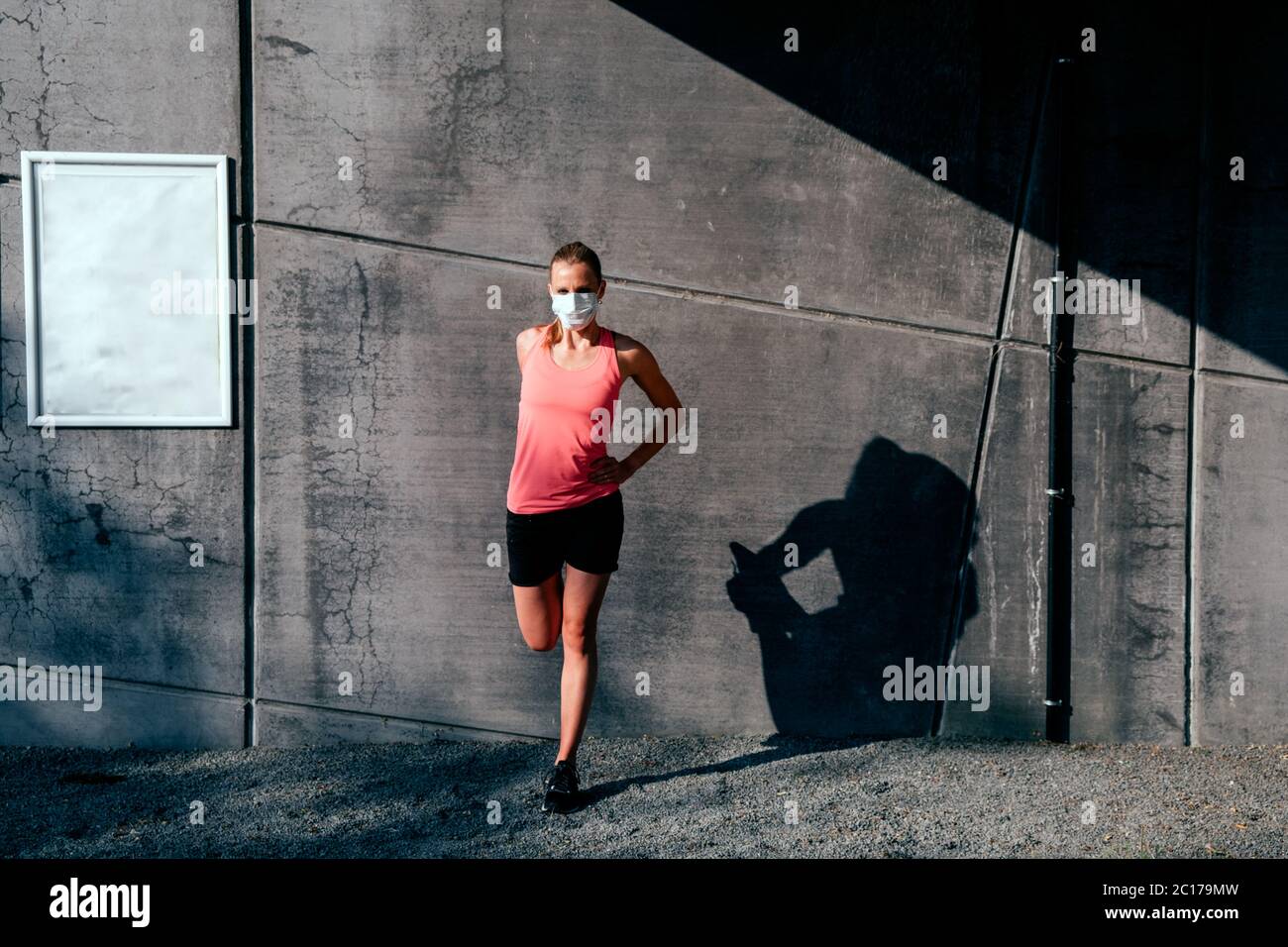 Gesunde kaukasische Frau in Sportkleidung mit Schutzmaske Stretching im Freien bei Sonnenuntergang auf einem Betonwand Hintergrund Stockfoto