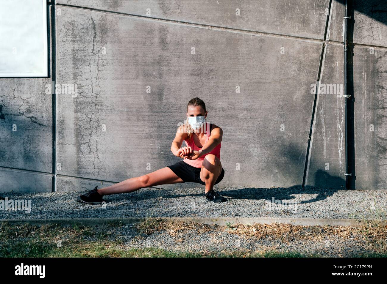 Gesunde kaukasische Frau in Sportkleidung mit Schutzmaske Stretching im Freien bei Sonnenuntergang auf einem Betonwand Hintergrund Stockfoto