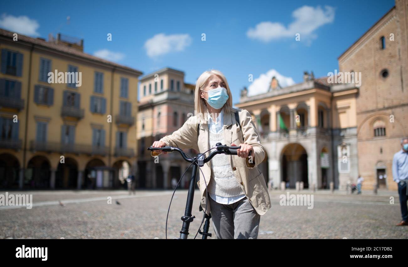 Reife maskierte Frau, die während einer Coronavirus-Pandemie ihr E-Bike in einer Stadt reitet Stockfoto