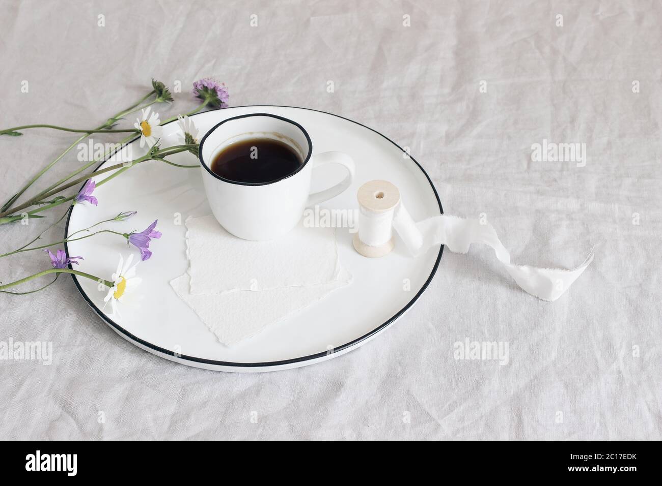 Frühling, Sommer Stillleben Szene. Unbeschriftete Grußkarten-Mockups aus Baumwollpapier auf Keramikplatte. Tasse Kaffee, Seidenband, Gänseblümchen und Bluebells Blumen Stockfoto