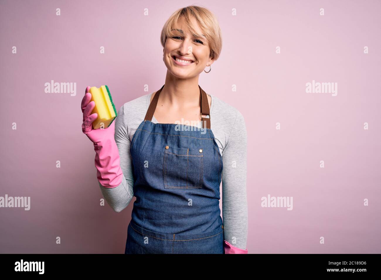 Junge blonde Putzfrau mit kurzen Haaren trägt Schürze und Handschuhe halten Peeling Geißel mit einem glücklichen Gesicht stehen und lächeln mit einem selbstbewussten smil Stockfoto