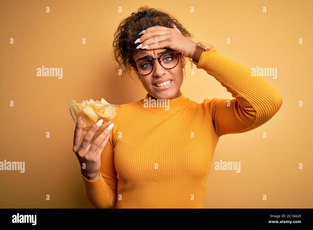 Junge afroamerikanische afro Mädchen hält Schüssel mit Kartoffeln Chips über gelbem Hintergrund betont mit Hand auf Kopf, schockiert mit Scham und Überraschung f Stockfoto