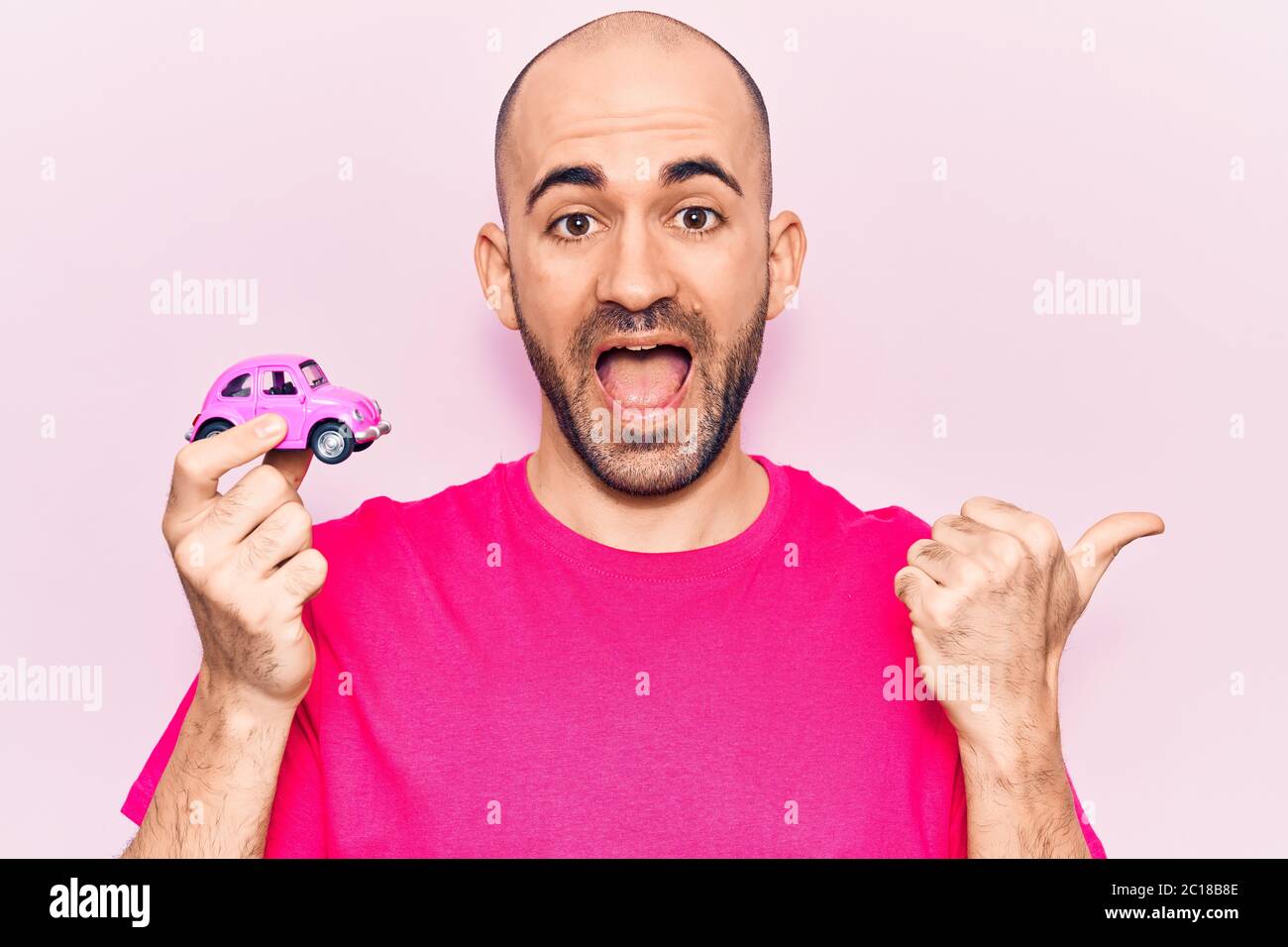 Junge gutaussehende Glatze Mann hält rosa kleinen Auto zeigt Daumen nach oben auf die Seite lächelnd glücklich mit offenem Mund Stockfoto
