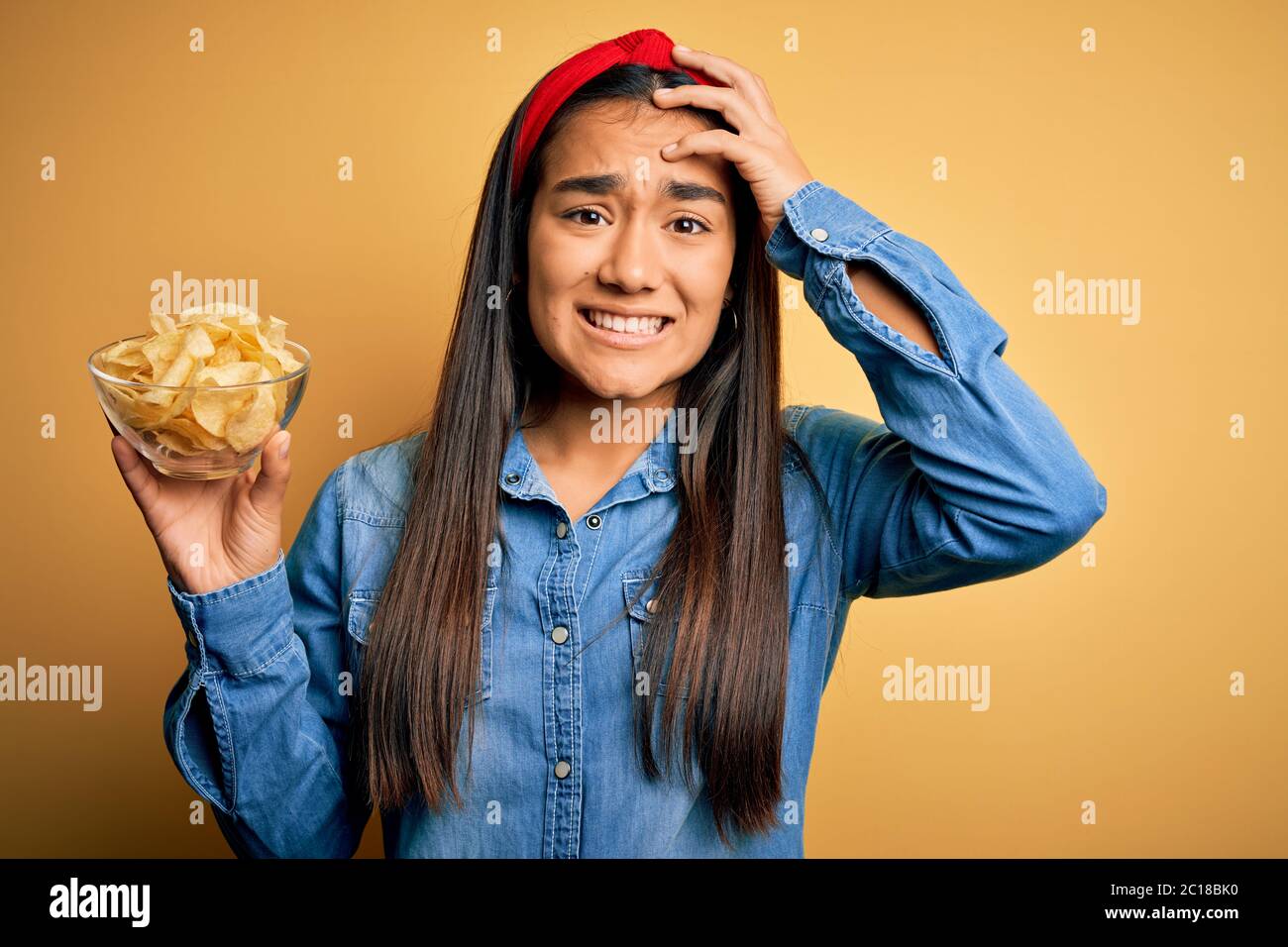 Junge schöne asiatische Frau hält Schüssel mit Chips Kartoffeln über isolierten gelben Hintergrund betont mit Hand auf Kopf, schockiert von Scham und Surpri Stockfoto
