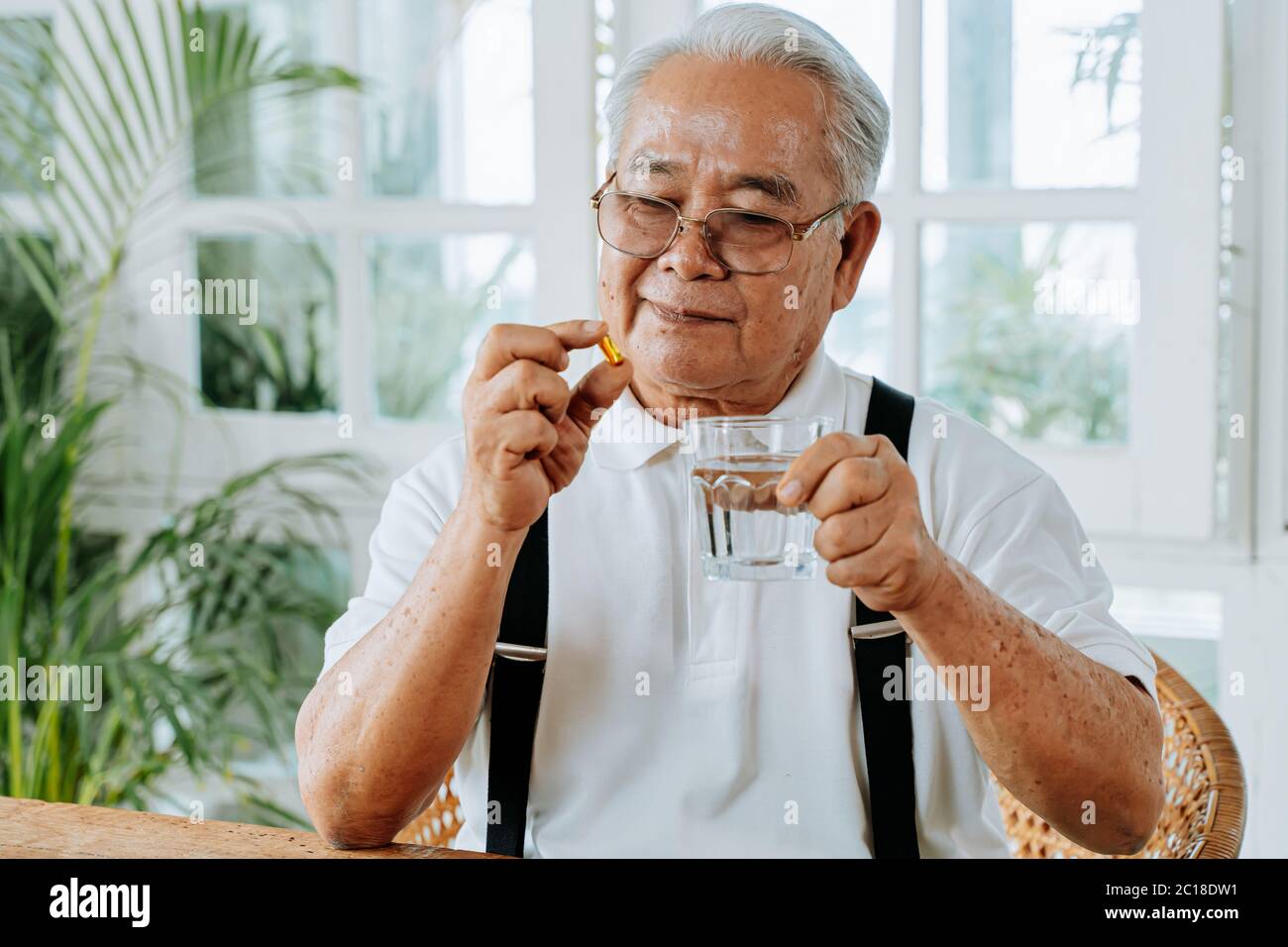 Senior Asian Mann mit Tasse Wasser und Pille Einnahme Heilmittel in gemütlichen Raum zu Hause. Alter Mann nimmt Medizin Stockfoto