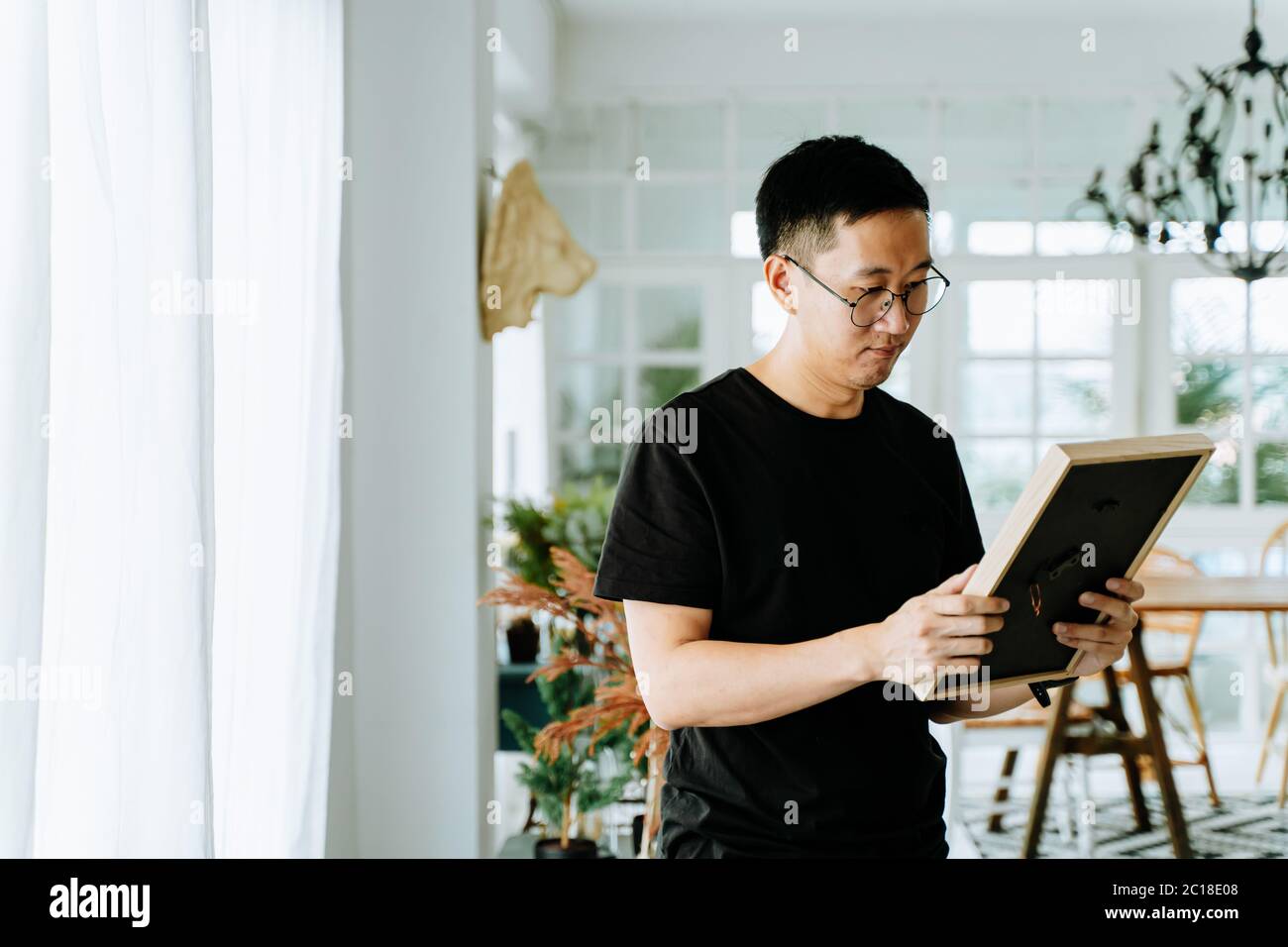 Traurige asiatische männlich in Brille Blick auf gerahmte Bild und Erinnerungen an die Lieben in gemütlichen Raum zu Hause Stockfoto