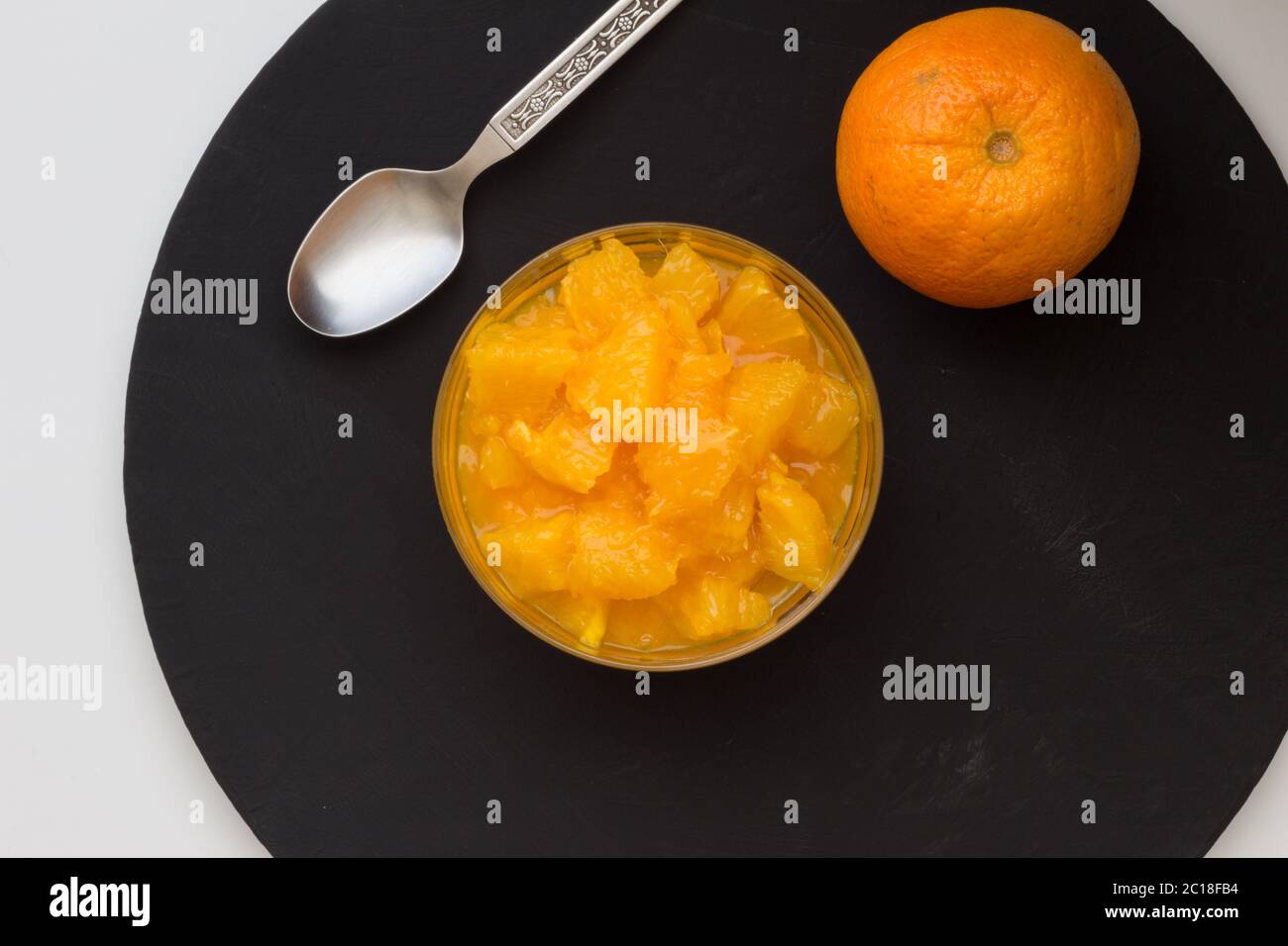 Orangenscheiben und Saft in Dessertschale auf schwarzem Rundschiefer mit ganzer Orange und Löffel. Draufsicht Stockfoto