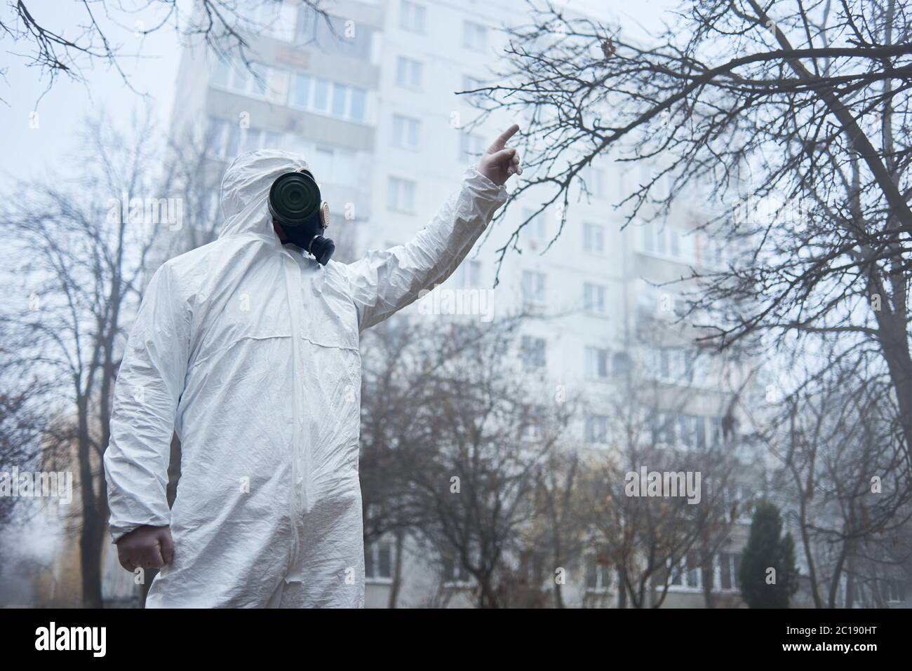 Wissenschaftler zeigt mit dem Finger auf, zwischen Herbstbäumen stehend. Mann in Gasmaske und Schutzkostüm posiert in leerer Stadt in der Nähe von Gebäuden. Konzept der städtischen Verschmutzung, feindliche Umwelt, Nebel. Stockfoto