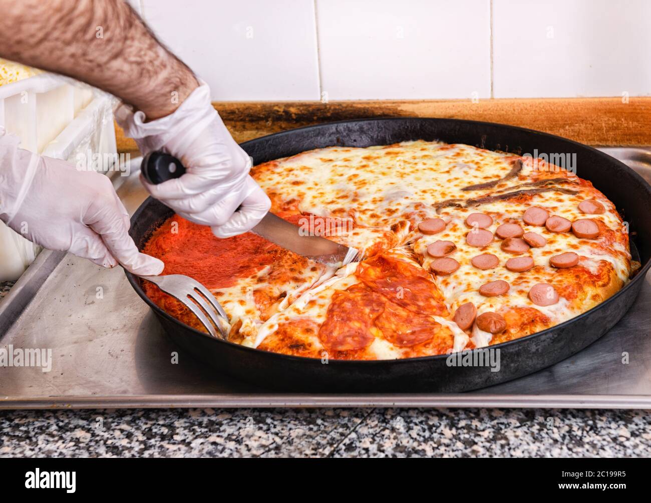 Der Koch schneidet eine italienische Pizza mit vier verschiedenen Belägen mit einem Messer und einer Gabel in einer Pizzeria-Küche in einer Nahaufnahme auf seinen Händen Stockfoto