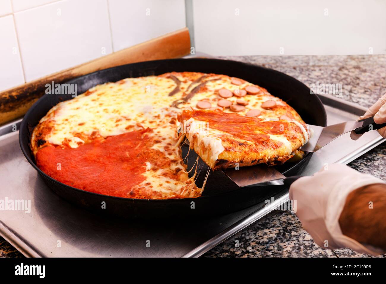 Der Koch nimmt eine Scheibe frisch gebackene Pizza aus einem Backblech und verwendet dabei einen Spatel aus nächster Nähe auf seine Handschlöcke Stockfoto