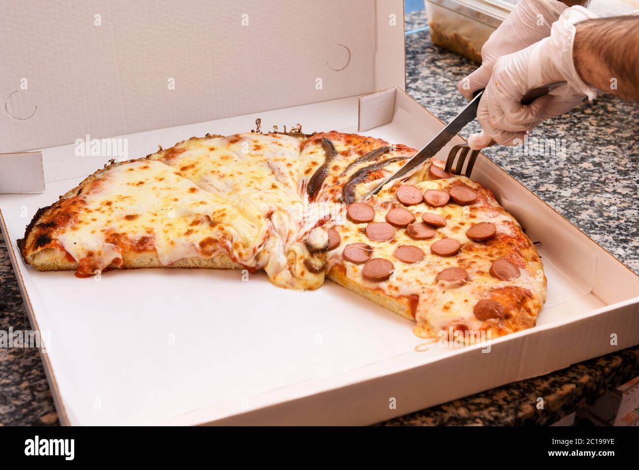 Koch mit Handschuhen schneidet eine Pizza mit verschiedenen Belägen in eine Pappschachtel zum Mitnehmen auf einer Granittheke in Nahaufnahme Stockfoto