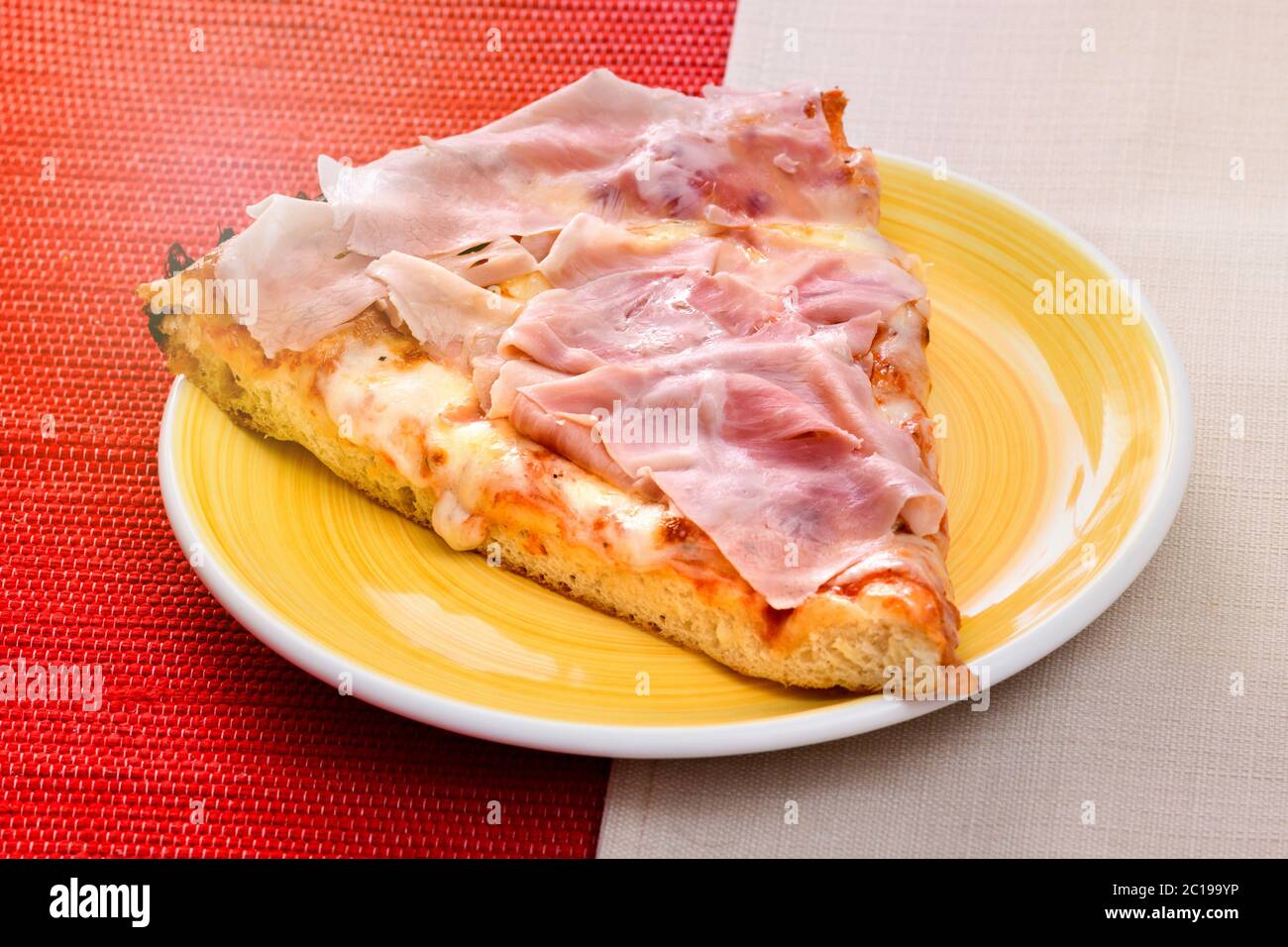 Italienische Pizza mit Schinken auf einer dicken Backbasis mit geschmolzenem Mozzarella und Tomatenmark auf einem gelben Teller auf einem zweifarbigen roten und serviert Stockfoto