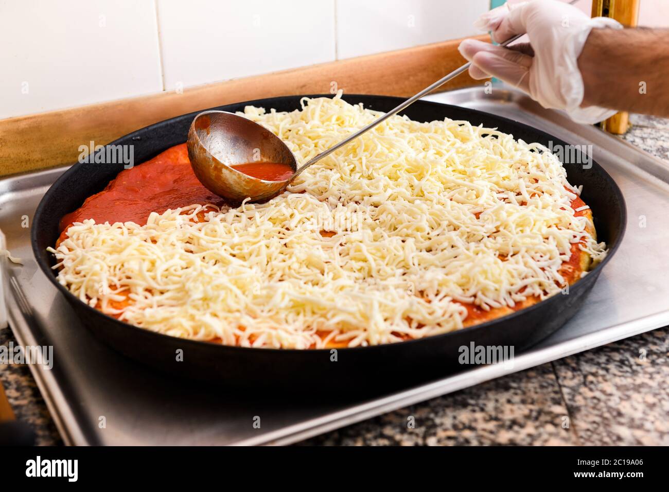 Der Koch löffelt Tomatenmark auf eine Pizzakruste, die mit Mozzarella-Käse in einer Pfanne bestreut wird, während er in einer Pizzeria zubereitet wird Stockfoto