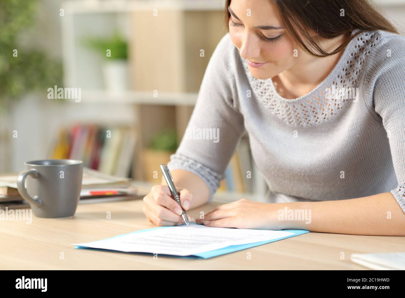 Glückliche Frau, die zu Hause auf einem Schreibtisch sitzt und den Vertrag liest und unterzeichnet Stockfoto