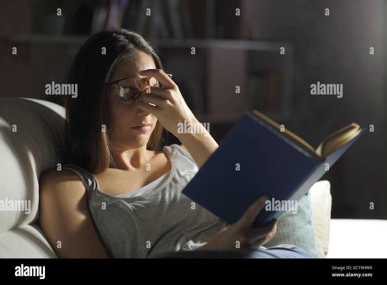 Frau mit Schmerzen leiden Augenbelastung Lesen eines Buches sitzt auf dem Sofa in der Nacht zu Hause Stockfoto