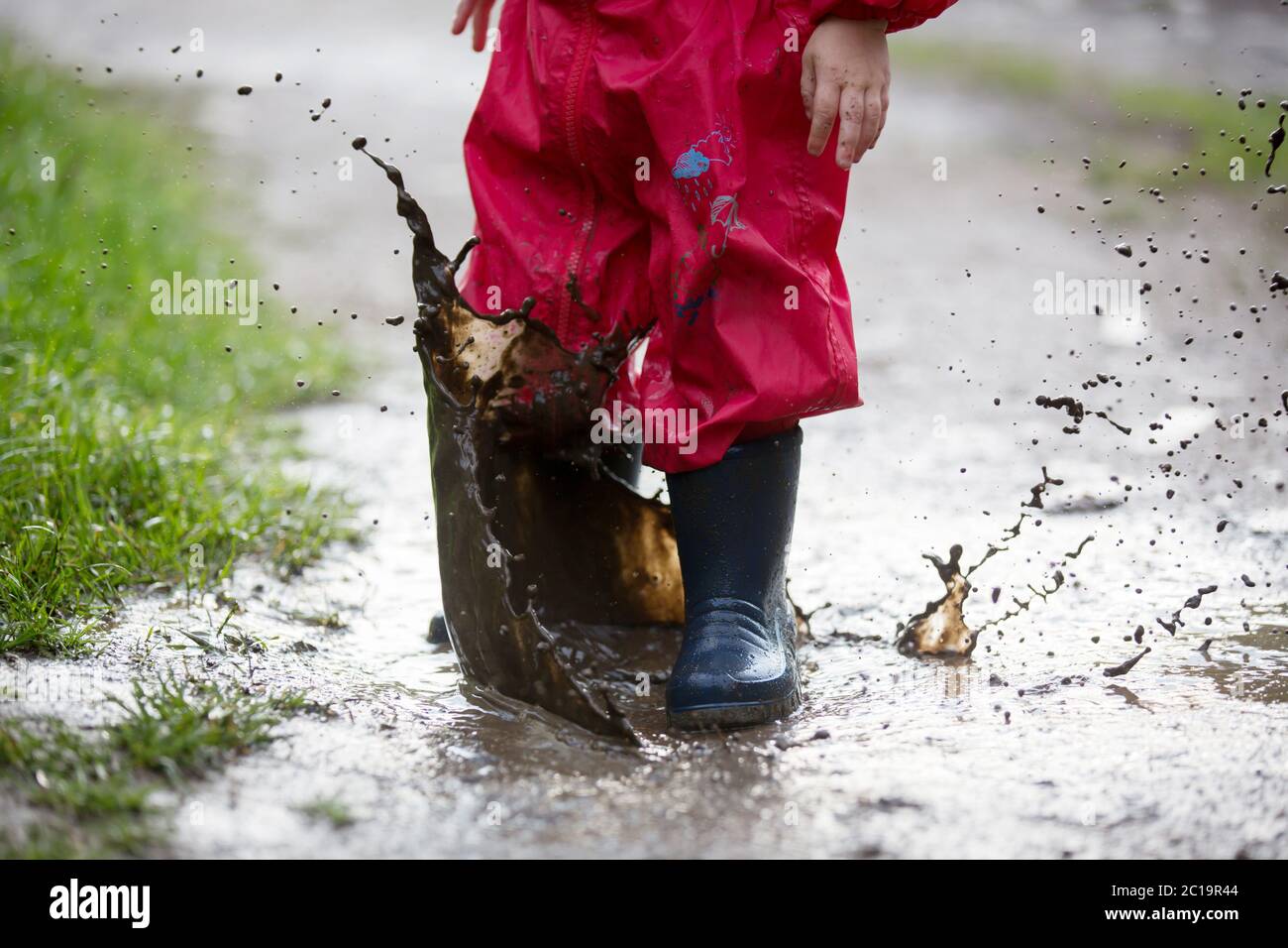 Süßer kleiner Kleinkind Junge, blondes Kind in rotem Regenmantel und blauen Stiefeln, im Regen in schlammigen Pfützen spielen, springen und lachen Stockfoto