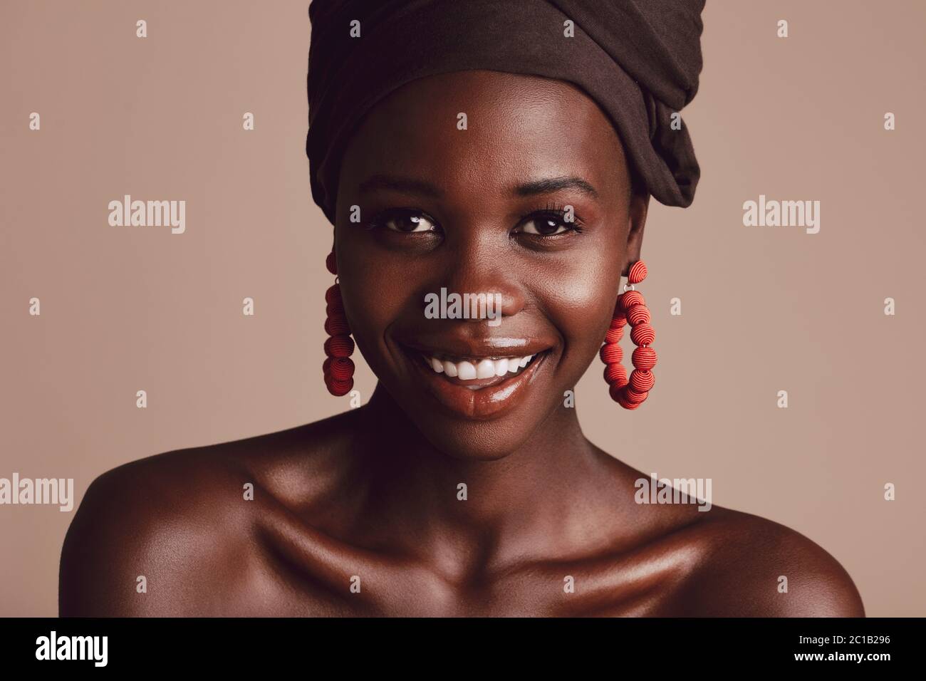 Nahaufnahme eines stilvollen afrikanischen weiblichen Modells mit Make-up Blick auf Kamera und lächelnd. Schöne Frau mit Turban vor beigem Hintergrund Stockfoto