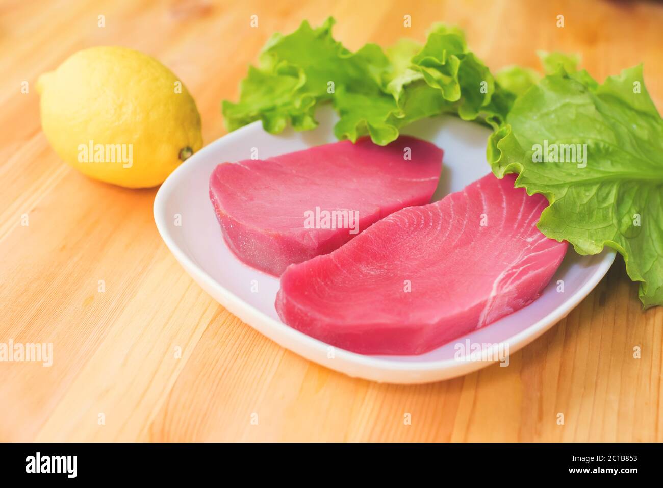 Zwei Stücke rohes frisches Thunfischfilet auf einem Teller mit großen Blättern grünen Salats und gelbe Zitrone auf einem Holztisch. Köstliche SE Stockfoto