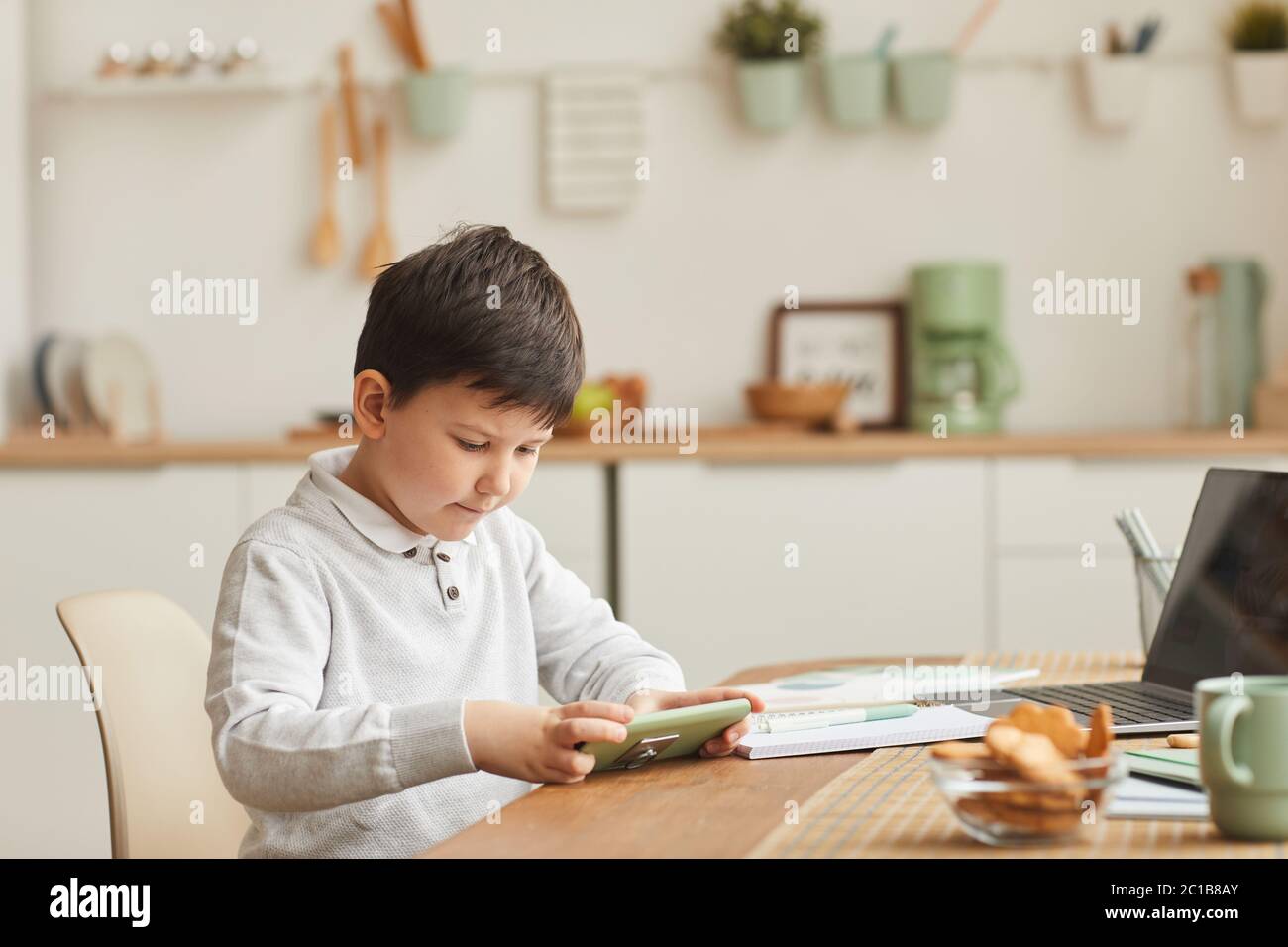 In warmen Farbtönen gehaltene Porträt eines niedlichen Jungen, der zu Hause unterwegs ist und am Schreibtisch mit Laptop in einer gemütlichen Küche sitzt, Platz zum Kopieren Stockfoto