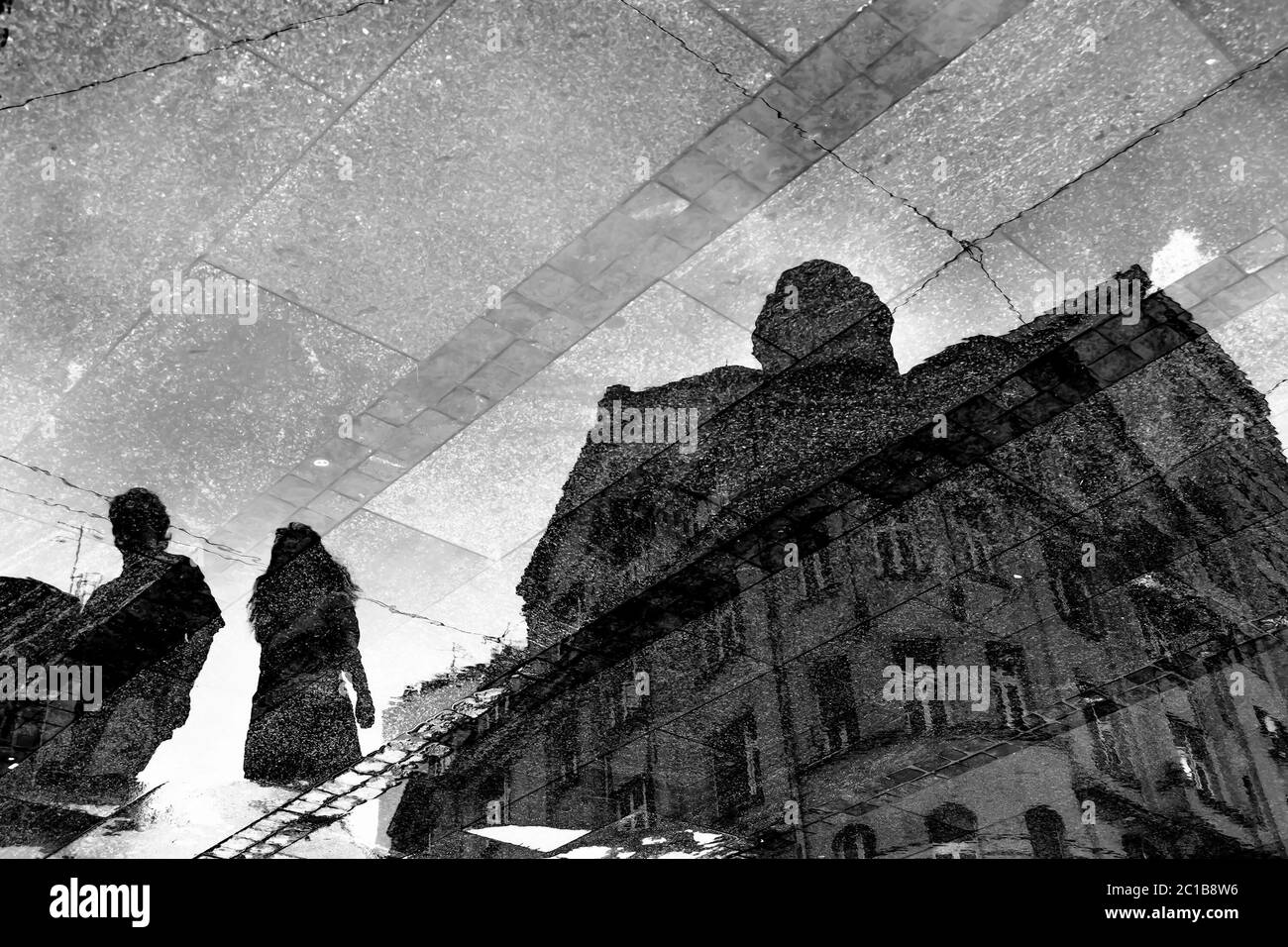 Verschwommene Spiegelschatten-Silhouetten in einer Pfütze eines neoklassizistischen Gebäudes und zwei Personen, die auf einer nassen Fußgängerzone in Schwarz und Weiß spazieren Stockfoto
