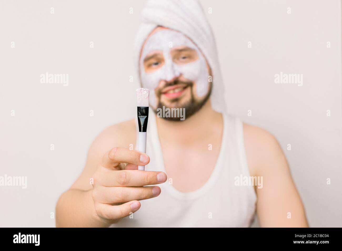 Jouful stattlich bärtigen Mann Anwendung Gesicht Schlammmaske auf Gesicht, zeigt kosmetische Bürste an der Kamera, isoliert auf weißem Hintergrund, Studio-Aufnahme. Konzentrieren Sie sich auf Stockfoto
