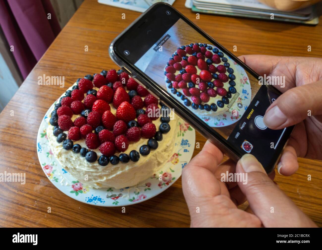 Eine Dame fotografiert mit einem Smartphone einen frisch gebackenen Kuchen, der mit Buttereier und weichen roten und violetten Sommerfrüchten verziert ist Stockfoto