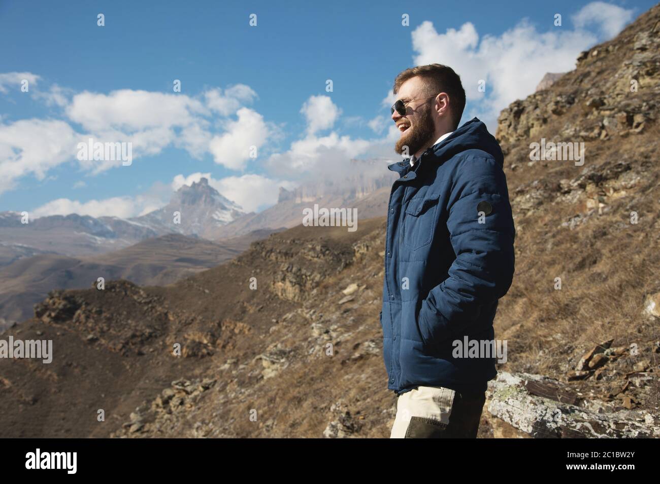 Hipster-Reisende in Daunenjacke und Sonnenbrille steht auf einem Berghang  vor der Kulisse epischer Felsen und lächelt. Die c Stockfotografie - Alamy