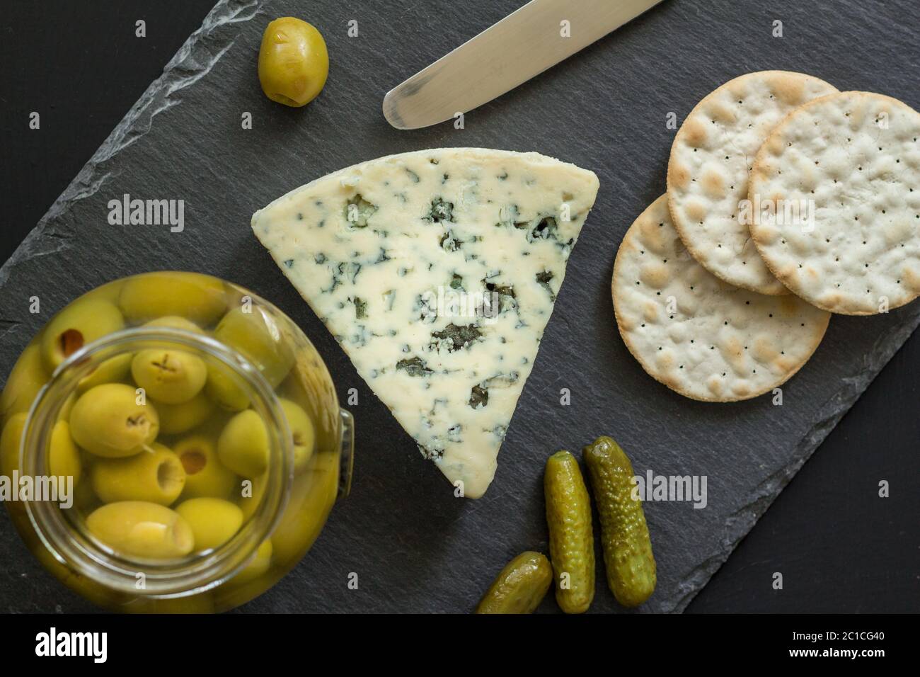 Käseplatte mit Blauschimmelkäse, Oliven, Gurken und Cracker auf schwarzem Schiefer - Foto von oben Stockfoto