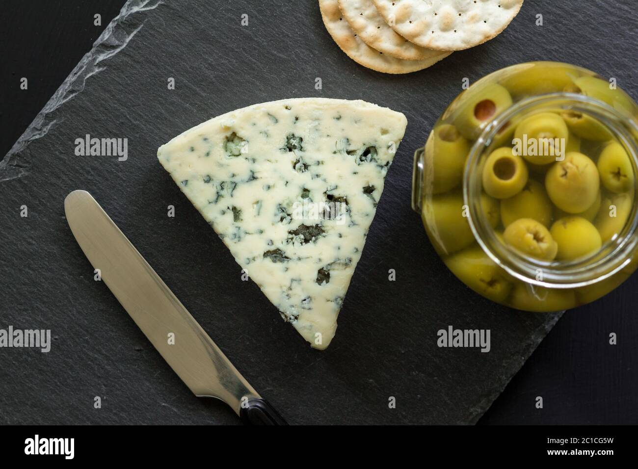Foto von Stilton Blue Cheese, Glas gefüllte Oliven und Cracker auf schwarzem Schiefer Käse Wildschwein Stockfoto