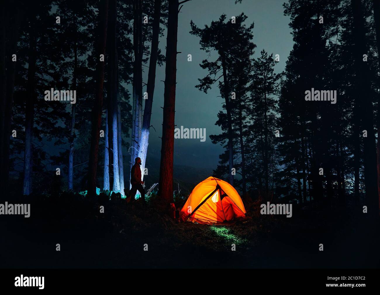 Bärtiger Mann Silhouette ist gegen Baumstamm im Nachtwald in der Nähe glühenden orange Zelt in den Bergen zu Fuß. Stockfoto