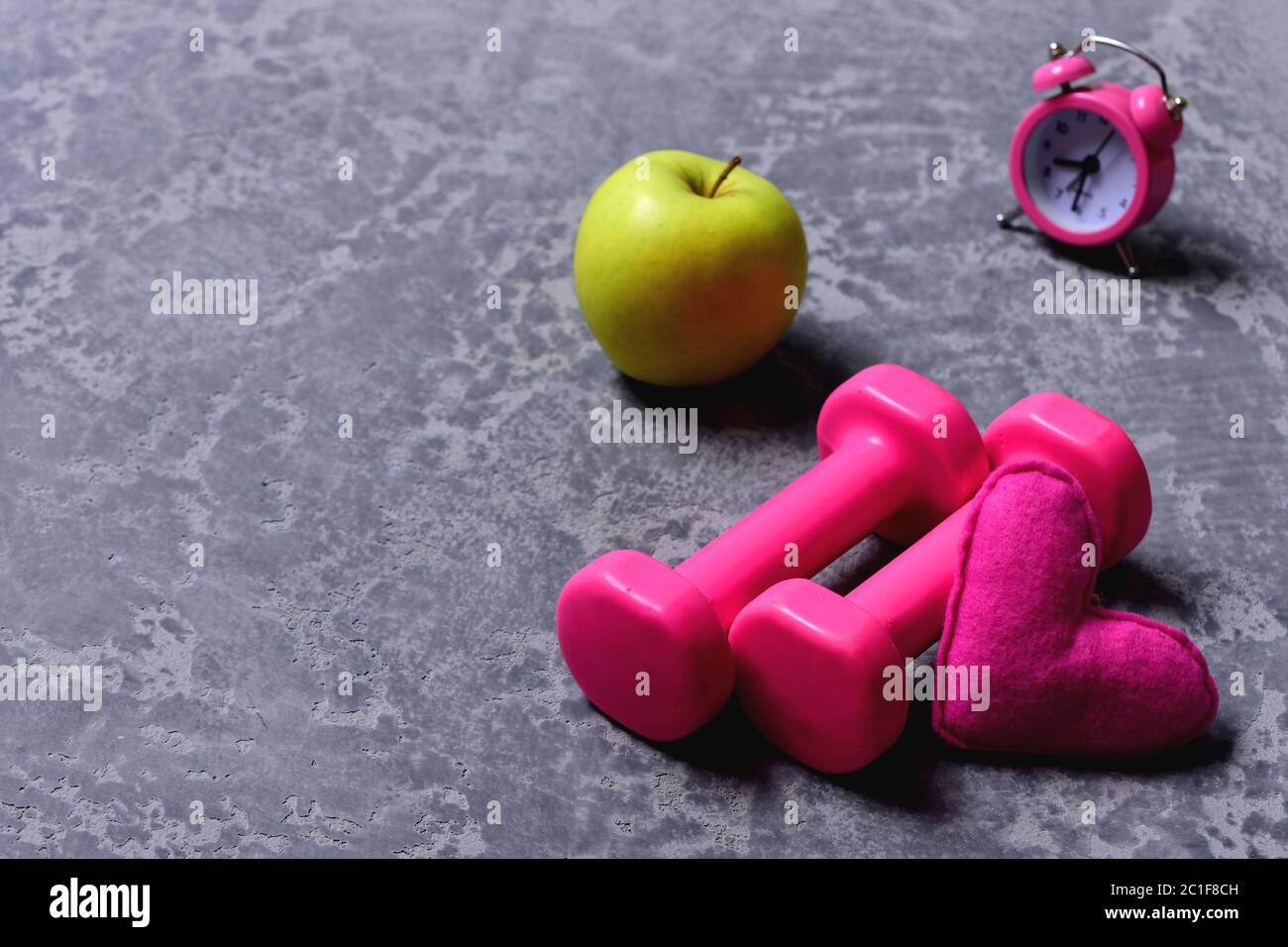 Liebe zum Sport und frühes Training. Damen-Style und Morgen-Workout-Konzept. Hanteln in rosa Farbe neben Apfel, Wecker und weiches Spielzeug Herz auf grauem Hintergrund. Herz, Apfel und Kunststoff-Langhanteln Stockfoto