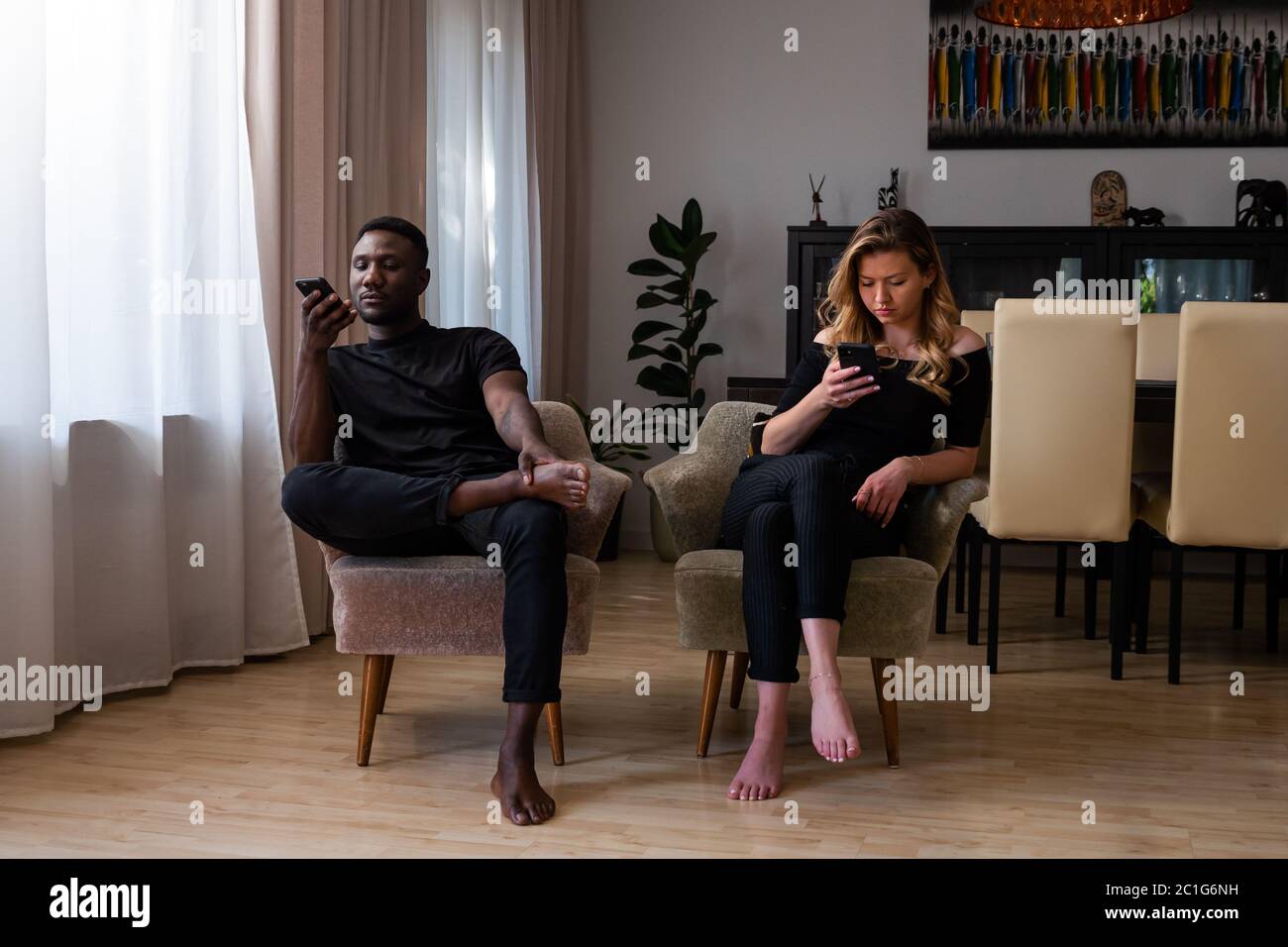 Junge gemischte Rennen Paar im Wohnzimmer sitzen Arme Stühle mit Smartphones weg suchen. Volle Länge. Stockfoto