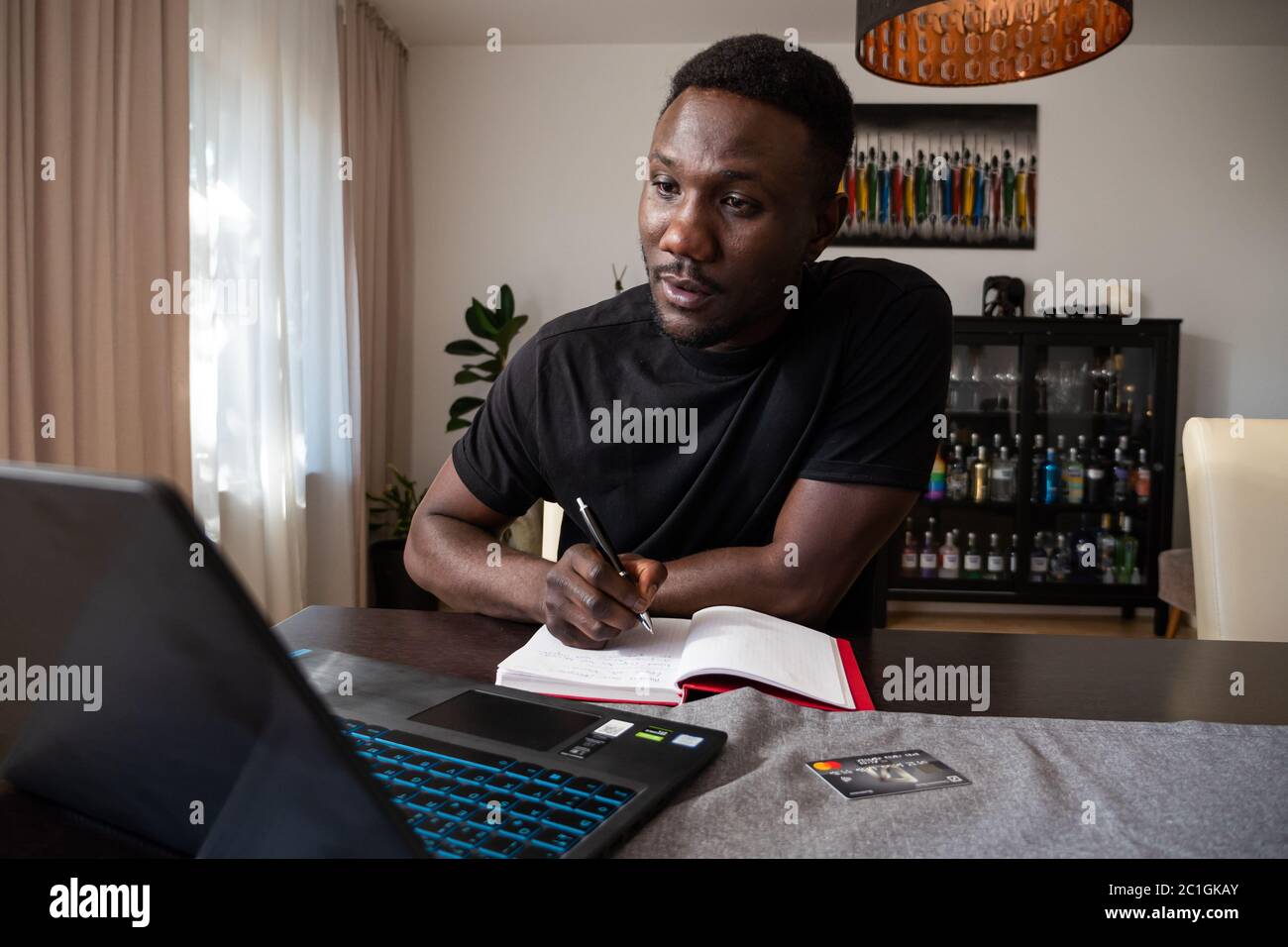 Junger schwarzer Mann, der im Wohnzimmer sitzende Schreibpapier und Schreibschriften von einem Laptop nimmt. Mittlere Aufnahme. Stockfoto
