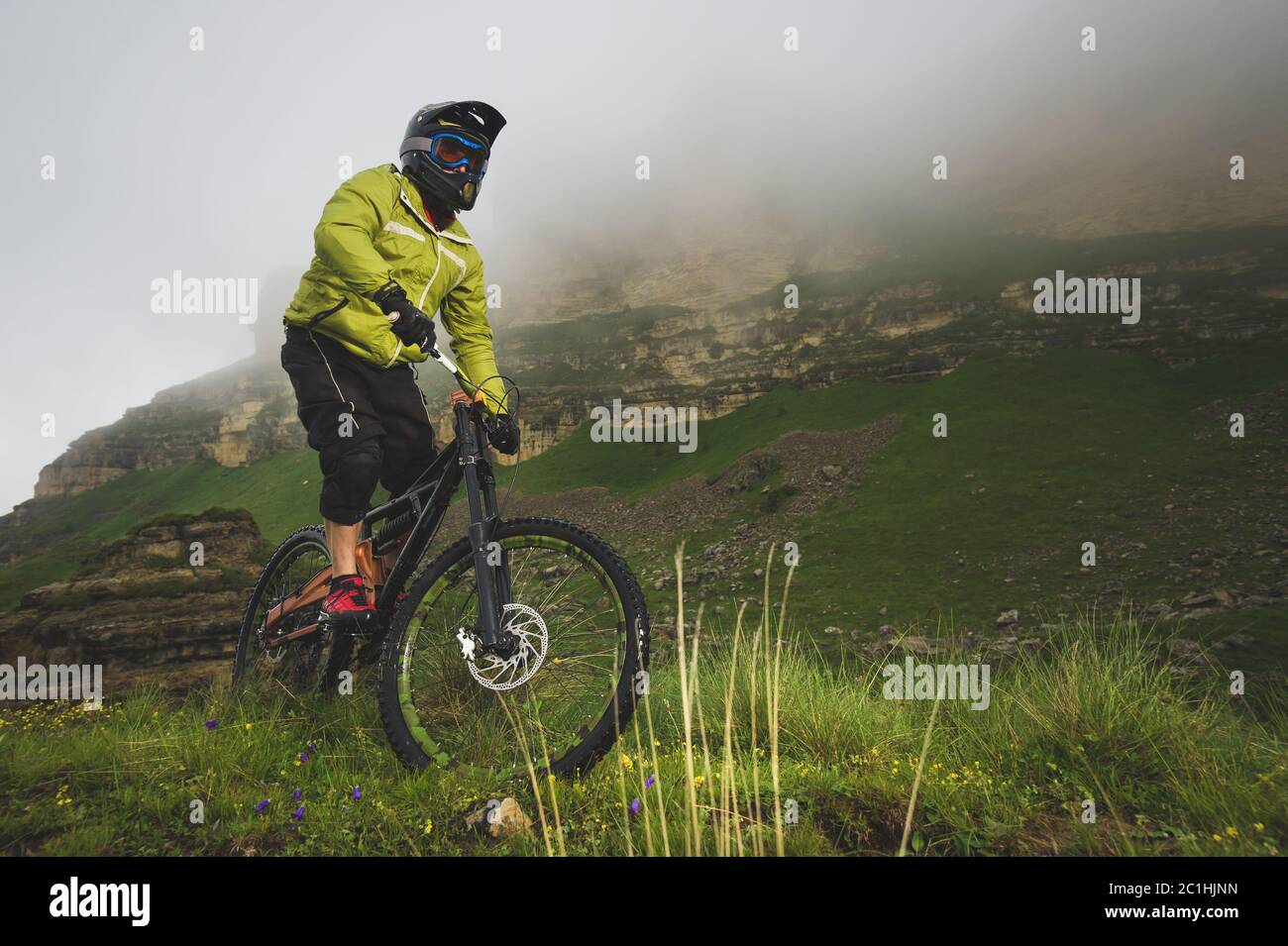 Ein erwachsener mtb-Radfahrer auf einem Mountainbike am Fuße einer Klippe, umgeben von grünem Gras. Niedrige Wolken. Nordkaukasus. Russland Stockfoto