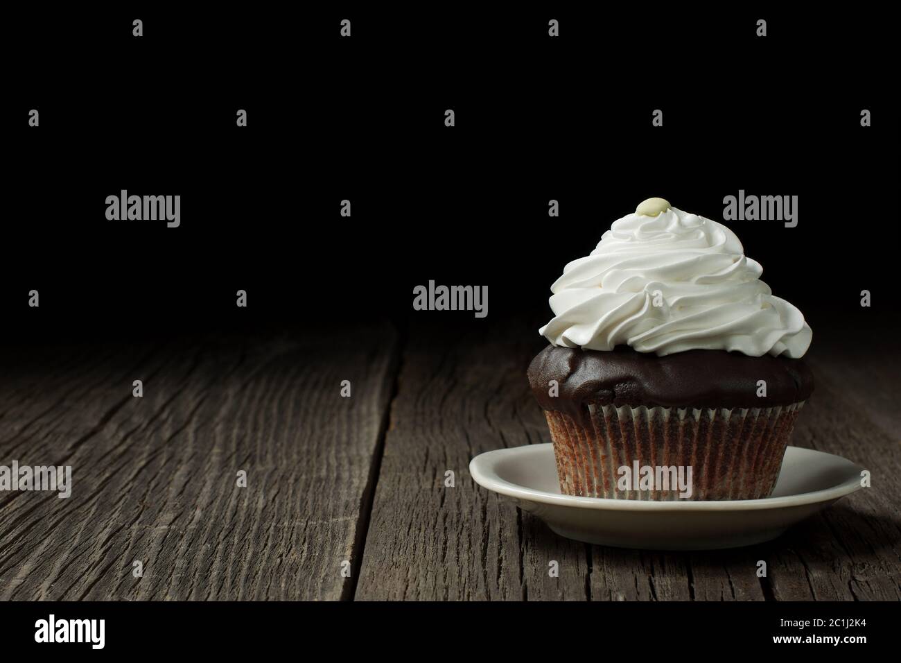 Ein Schokoladenkuchen in einem typischen geriffelten Cupcake Liner, mit Schlagsahne gekrönt Er steht auf einer kleinen Keramikuntertasse, auf verwitterten Holzplanken. Stockfoto