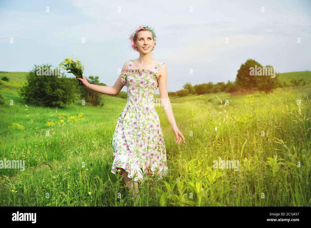 Junge glücklich lächelndes Mädchen in einem Calico Kleid mit einem Blumenstrauß ist entlang einer Landstraße mit grünem Gras zu Fuß Stockfoto