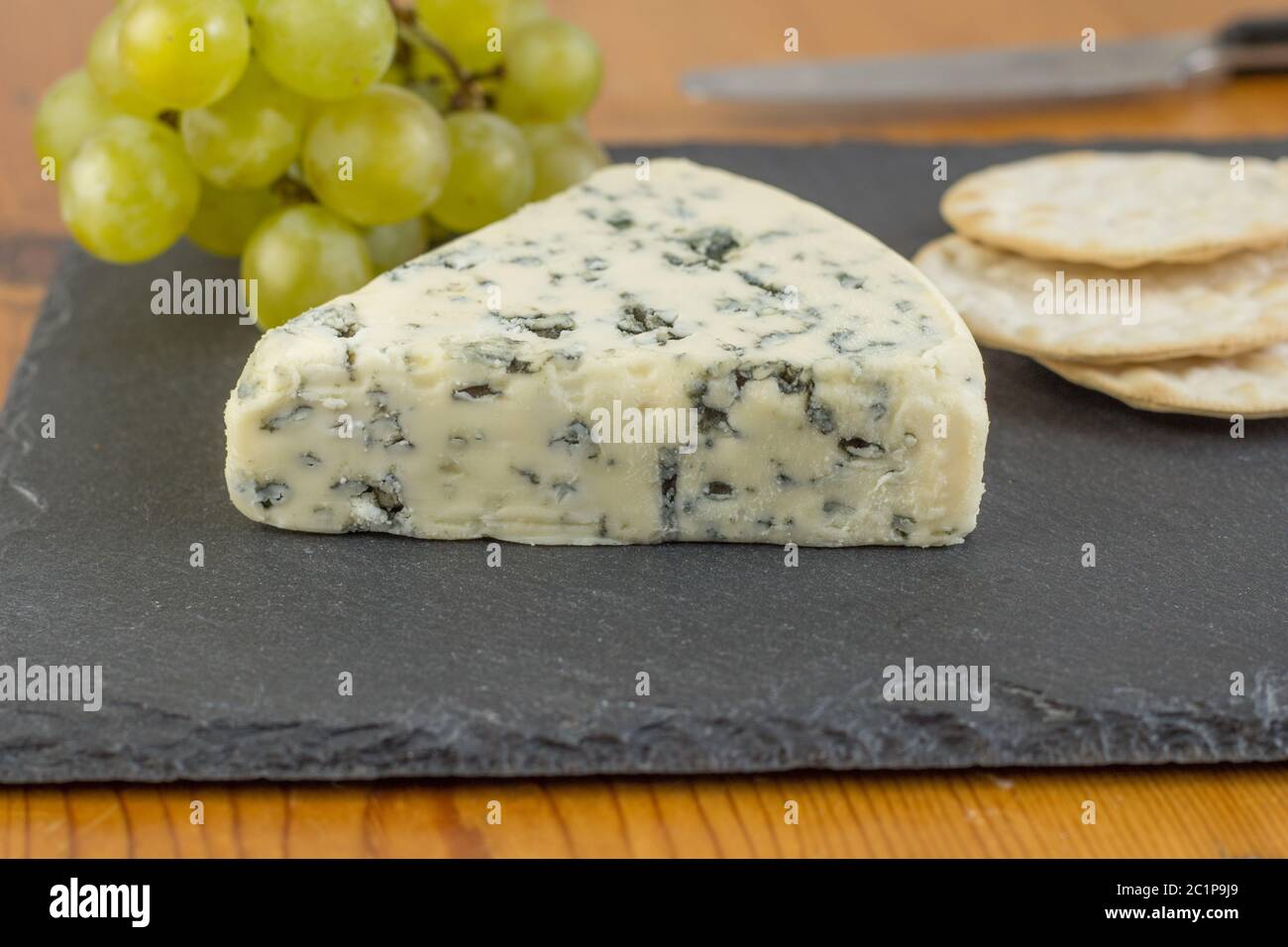 Blue Cheese wedge auf schiefer board schließen oben mit Defokussierten grüne Weintrauben und Cracker im Hintergrund Stockfoto