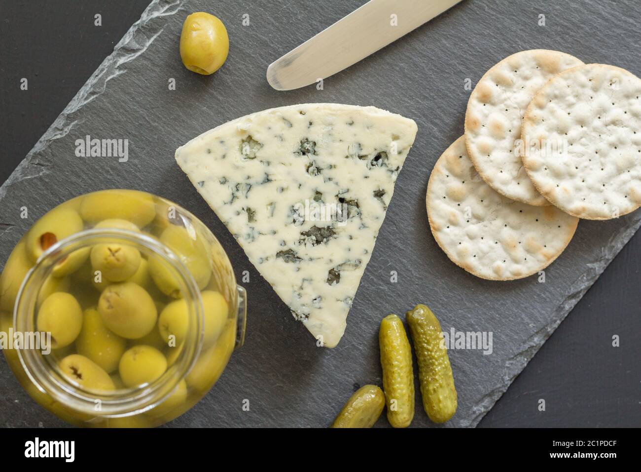 Blauer Käsekeil mit Crackern und Glas grüner Oliven auf schwarzem Schiefer Käseplatte isoliert Stockfoto